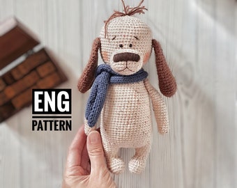 May include: Crocheted dog toy with brown ears, a blue scarf, and a white body. The toy is sitting on a white wooden surface. ENG PATTERN is written in black text on a white background.