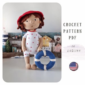 Puede incluir: Una muñeca de ganchillo con un sombrero rojo y un vestido blanco con lunares rojos, sosteniendo un salvavidas azul y blanco. La muñeca tiene el pelo rizado castaño y está de pie frente a una escena costera en miniatura. El texto dice "CROCHET PATTERN PDF IN ENGLISH".