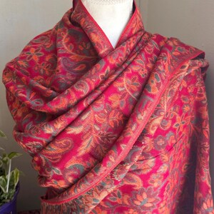May include: A vibrant red shawl with an intricate floral paisley pattern in shades of orange, green, and purple. The shawl is draped over a mannequin, showcasing its flowing design and detailed border. This textile accessory adds a touch of elegance.