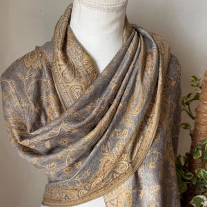Op de afbeelding: Een grijze en gouden paisley-patroon sjaal gedrapeerd over een mannequin. De sjaal heeft een gedetailleerd bloemmotief met een gouden rand. De stof lijkt zacht en vloeiend, geschikt voor een sjaal of omslagdoek.