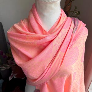 Pode incluir: Lenço com estampa paisley em tons de pêssego e rosa, drapeado sobre um manequim. O lenço tem uma textura macia e fluida e apresenta um design detalhado. A paleta de cores inclui tons de pêssego, rosa e um toque de roxo.