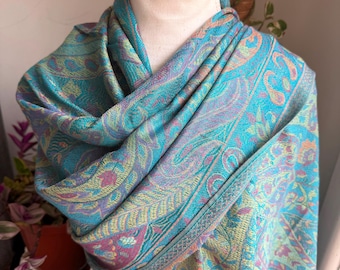 May include: A teal paisley patterned scarf draped over a mannequin. The scarf features a detailed design with shades of teal, purple, and yellow. The edges are finished with a subtle fringe. The scarf is made of a soft, flowing fabric.