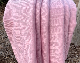 May include: A soft pink blanket with a woven herringbone pattern. The blanket is draped over a wooden post, showcasing its texture and color. The fabric appears to be lightweight and suitable for a throw or decorative piece.