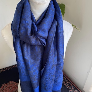 May include: A blue scarf with a paisley pattern is draped over a mannequin. The scarf has a dark blue border and fringe. The paisley pattern is a lighter shade of blue. The scarf is made of a soft, flowing fabric.