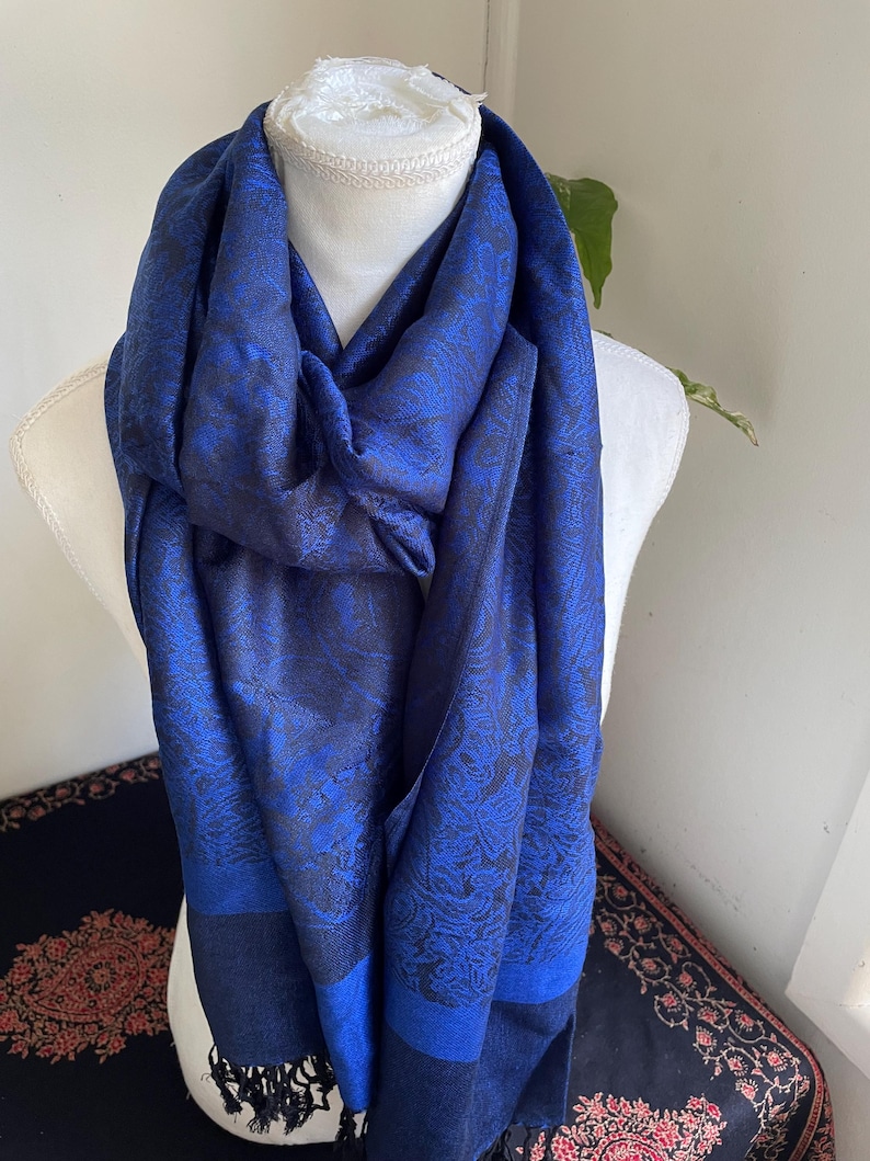 May include: A blue scarf with a paisley pattern is draped over a mannequin. The scarf has a dark blue border and fringe. The paisley pattern is a lighter shade of blue. The scarf is made of a soft, flowing fabric.