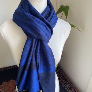 May include: A blue scarf with a floral pattern and black fringe is draped over a white mannequin. The scarf has a dark blue border and a black and blue floral design. The scarf is made of a soft material.