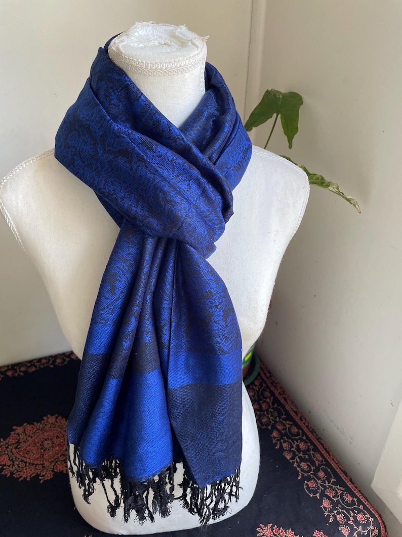 May include: A blue scarf with a floral pattern and black fringe is draped over a white mannequin. The scarf has a dark blue border and a black and blue floral design. The scarf is made of a soft material.