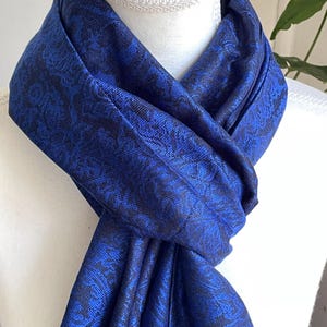 May include: A royal blue scarf with a dark blue floral pattern is draped over a white mannequin. The scarf is made of a soft, flowing fabric and is knotted at the front. The scarf is a fashion accessory.