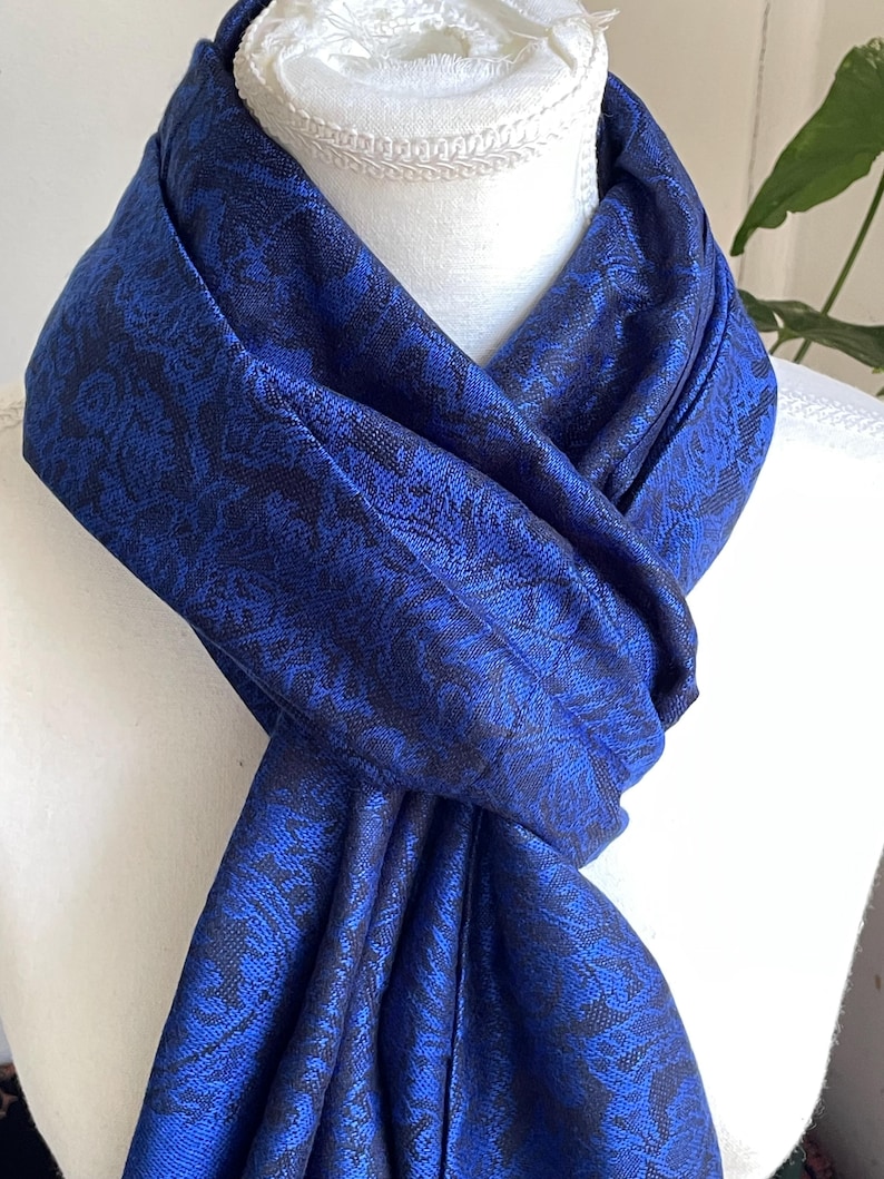 May include: A royal blue scarf with a dark blue floral pattern is draped over a white mannequin. The scarf is made of a soft, flowing fabric and is knotted at the front. The scarf is a fashion accessory.