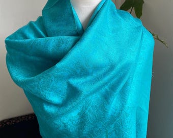 May include: A vibrant turquoise shawl draped over a mannequin. The shawl features a subtle paisley pattern and is made of a soft, flowing fabric. The bright color and delicate design make it a stylish accessory.
