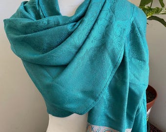 May include: A teal-colored scarf draped over a mannequin. The scarf has a subtle paisley pattern and a decorative border with a detailed design in shades of blue, orange, and cream. The scarf is made of a soft, flowing fabric.