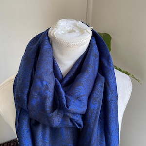 May include: A royal blue scarf with a dark floral pattern is draped over a white mannequin. The scarf's fabric has a subtle sheen, and the floral design adds a touch of elegance. The scarf is a versatile accessory.