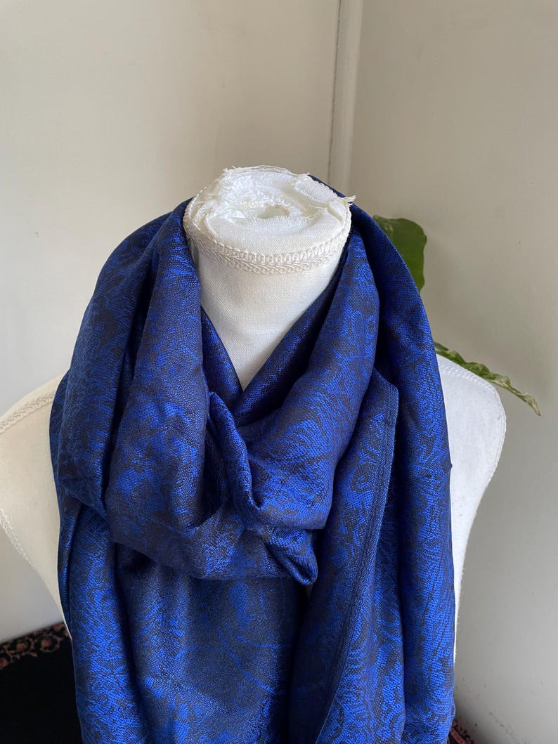 May include: A royal blue scarf with a dark floral pattern is draped over a white mannequin. The scarf's fabric has a subtle sheen, and the floral design adds a touch of elegance. The scarf is a versatile accessory.