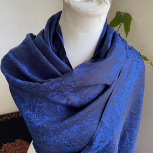 May include: A draped, blue scarf with a paisley pattern. The scarf is displayed on a white mannequin head. The scarf's color is a deep blue with a darker blue paisley design. The fabric appears soft and flowing.