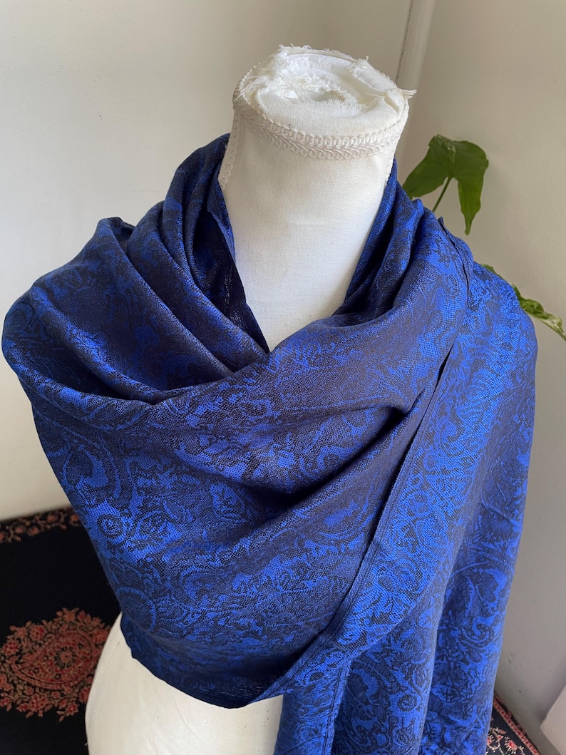 May include: A draped, blue scarf with a paisley pattern. The scarf is displayed on a white mannequin head. The scarf's color is a deep blue with a darker blue paisley design. The fabric appears soft and flowing.