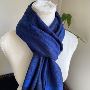 May include: A royal blue scarf with a black floral pattern is draped over a white mannequin. The scarf is made of a soft fabric and has a subtle sheen. The scarf is a fashion accessory.