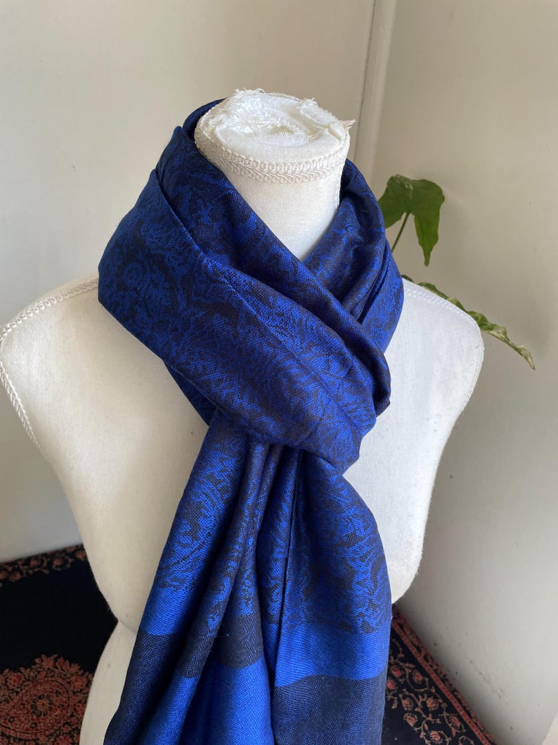 May include: A royal blue scarf with a black floral pattern is draped over a white mannequin. The scarf is made of a soft fabric and has a subtle sheen. The scarf is a fashion accessory.