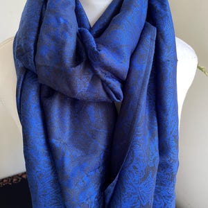 May include: A vibrant blue scarf with a detailed floral pattern. The scarf is draped over a mannequin, showcasing its texture and design. The fabric appears to have a subtle sheen, enhancing the rich color and intricate details.
