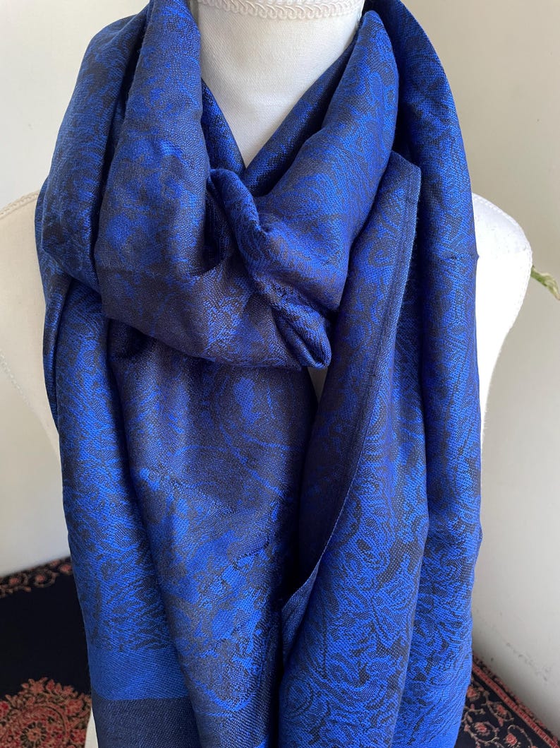 May include: A vibrant blue scarf with a detailed floral pattern. The scarf is draped over a mannequin, showcasing its texture and design. The fabric appears to have a subtle sheen, enhancing the rich color and intricate details.