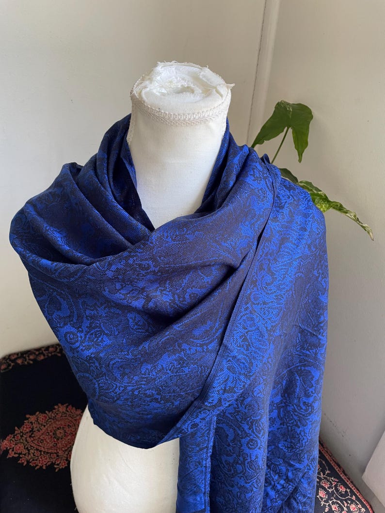 May include: A draped, royal blue shawl with a paisley pattern. The fabric is a deep blue with darker blue detailing, creating a textured appearance. The shawl is displayed on a white mannequin head.