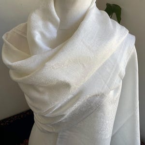 May include: A cream-coloured scarf draped over a mannequin. The scarf features a subtle paisley pattern and a fringed edge. The fabric appears soft and lightweight, suitable for layering.