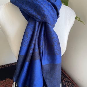 May include: A royal blue scarf with a black floral pattern and fringe. The scarf is draped over a white mannequin. The scarf has a dark blue border and black fringe at the bottom. The scarf is made of a soft material.