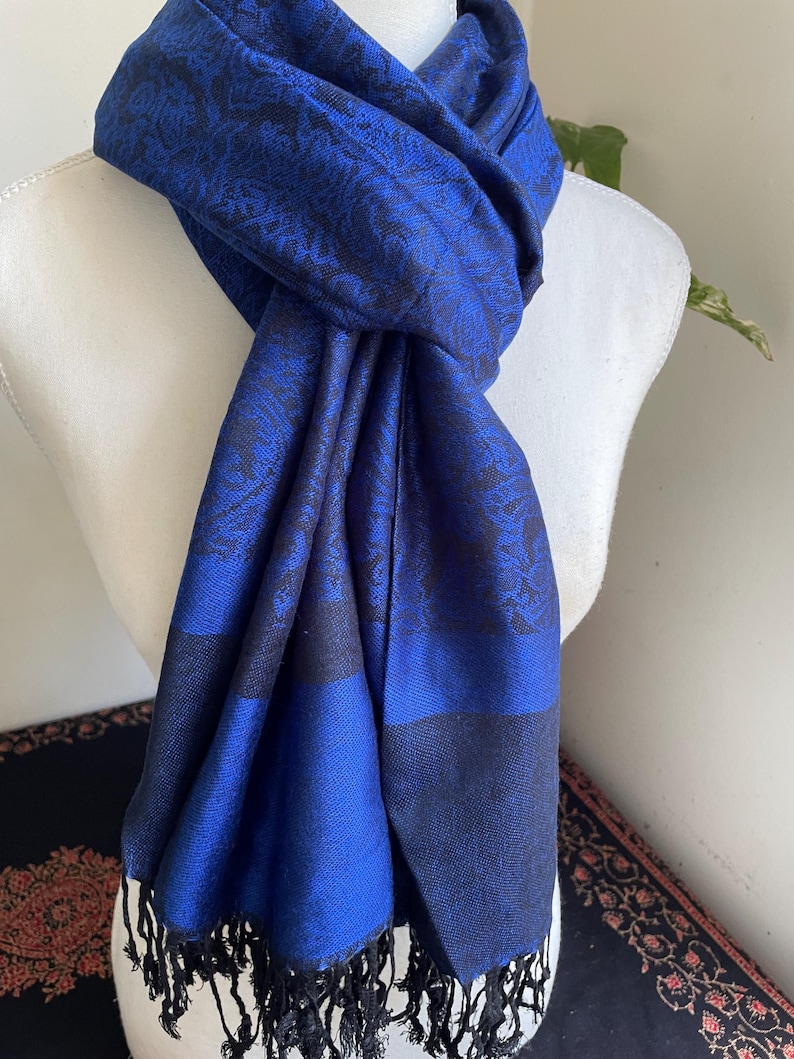 May include: A royal blue scarf with a black floral pattern and fringe. The scarf is draped over a white mannequin. The scarf has a dark blue border and black fringe at the bottom. The scarf is made of a soft material.