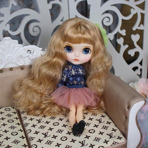 May include: A doll with long blonde hair wearing a blue and white sweater and a pink tulle skirt. The doll is sitting on a sofa with a brown and white patterned fabric.