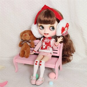 Pode incluir: Uma boneca com cabelo castanho usando uma faixa de cabeça vermelha e branca e um vestido rosa e vermelho está sentada em um banco rosa com um urso de pelúcia marrom.