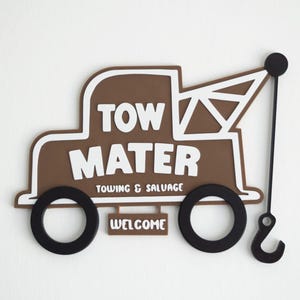 Può includere: Un'insegna da parete marrone e bianca a forma di carro attrezzi con il testo TOW MATER, TOWING & SALVAGE e WELCOME. L'oggetto presenta due ruote circolari nere e un braccio della gru nero con un gancio che si estende sul lato destro.