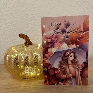 Könnte beinhalten: Ein goldener Glaskürbis mit Innenbeleuchtung neben einer rechteckigen Planereinlage mit dem Text HERBST Jahreschallenge, einer Illustration einer Person mit langem welligem Haar und Regenschirm sowie herbstlichen Blumen im Hintergrund.
