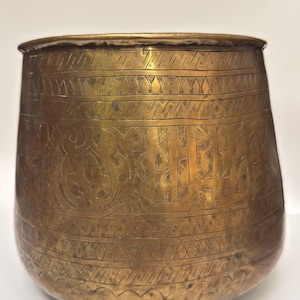 Peut inclure: Un bol en laiton avec une base arrondie et une large ouverture. La surface extérieure présente des motifs gravés à la main, notamment des bandes horizontales de lignes diagonales, des motifs triangulaires et une calligraphie centrale.