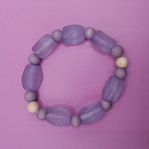 May include: A lavender beaded bracelet with a mix of shapes and colors. The bracelet features large, rectangular beads and smaller, round beads in shades of lavender and a single cream-colored bead. The bracelet is set against a solid lavender background.