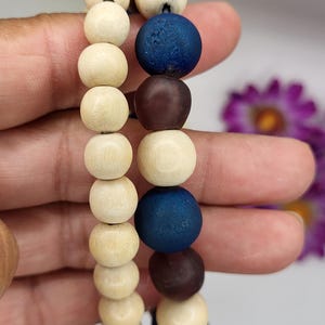 May include: Two beaded bracelets. One bracelet features light wood beads. The other bracelet has a mix of dark blue, brown, and light wood beads. The bracelets are held in a hand against a soft focus background.