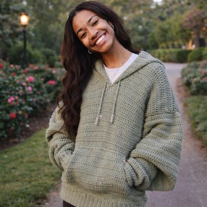 May include: A sage green crochet hoodie with a drawstring hood and front pocket. The handmade sweater has long sleeves and a relaxed fit. The person wearing the hoodie is smiling in an outdoor setting.