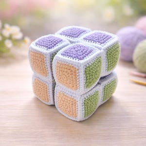 Può includere: Un cubo puzzle all'uncinetto fatto a mano. Il cubo è composto da quadrati individuali nei toni del viola, verde e pesca, con bordi bianchi. Il cubo è su una superficie di legno chiaro. Dimensioni: circa 8 cm x 8 cm x 8 cm.