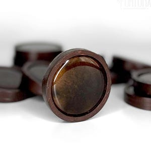 May include: Close-up of several brown, circular game pieces with a dark, glossy center. The pieces have a dark brown, leather-like border. The background is white, with the top right corner showing the text "GAMMON FORTUNA".