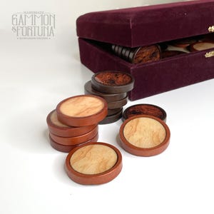 Puede incluir: Un juego de backgammon hecho a mano con piezas de madera. Las piezas son de forma ovalada con un centro de madera clara y un borde marrón oscuro. El juego incluye un estuche de terciopelo burdeos. El texto "GAMMON & FORTUNA" es visible.