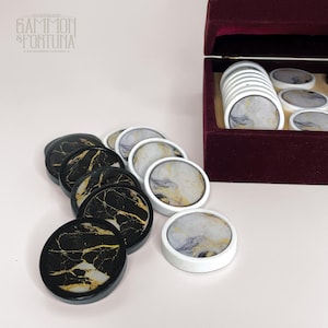 May include: A set of backgammon checkers in black and white with gold veining, and white with gray and gold marbling. The checkers are displayed with a burgundy velvet box. The brand name "GAMMON & FORTUNA" is visible.