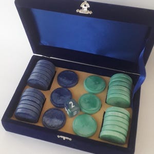 May include: A navy blue velvet-lined box containing two stacks of dark blue circular game pieces, two stacks of light green circular game pieces, three loose dark blue pieces, three loose light green pieces, and one dark blue eight-sided die.