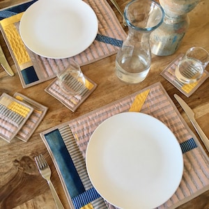 Op de afbeelding: Een tafelsetting met twee witte borden op placemats met patronen. De placemats hebben een patchwork-ontwerp met gele, blauwe en grijze strepen. Onderzetters, glazen, een waterkan en bestek maken de opstelling compleet.