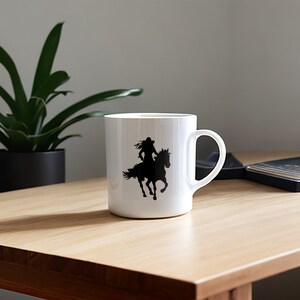 May include: White ceramic mug featuring a black silhouette of a person riding a horse. The design shows a person wearing a hat and flowing hair. The mug has a curved handle and is set on a wooden surface.