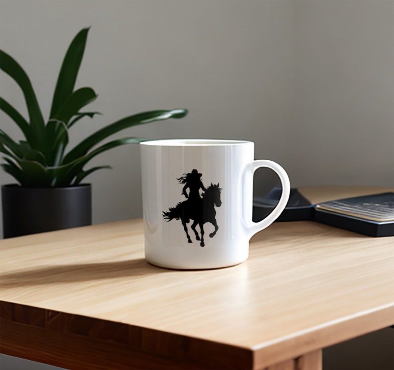 May include: White ceramic mug featuring a black silhouette of a person riding a horse. The design shows a person wearing a hat and flowing hair. The mug has a curved handle and is set on a wooden surface.