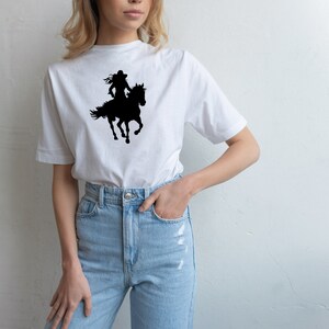 May include: White t-shirt featuring a black silhouette of a person riding a horse. The rider is wearing a cowboy hat. The shirt is paired with light blue jeans. The overall theme is Western and equestrian.