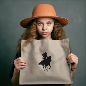 May include: A tan tote bag featuring a black silhouette of a person riding a horse. The bag is held in front of a person wearing an orange hat. The background is a solid gray color.