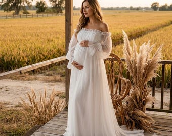 May include: A floor-length white maternity gown featuring an off-the-shoulder neckline with a ruffled trim, long sheer bishop sleeves with gathered cuffs, and a flowing skirt made of lightweight, semi-sheer fabric that drapes to the floor.