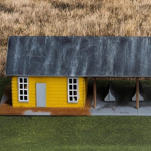 May include: A miniature yellow building with a gray roof and a covered area. The building has white-framed windows and a gray door. The structure is set on a brown base, with a green and tan background.