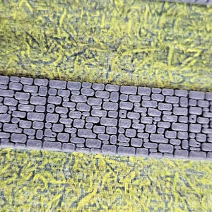 May include: Close-up of a miniature stone wall with a textured surface, composed of various-sized gray stones. The wall is set against a backdrop of artificial green grass, creating a rustic, textured appearance.