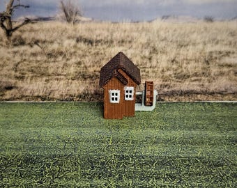 May include: A miniature wooden house with a brown roof and white windows. The house has a small water wheel attached to the side. The house is sitting on a green grassy surface.