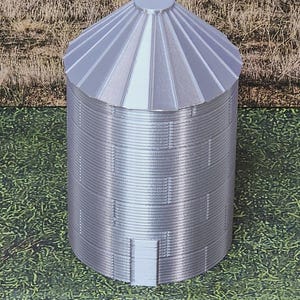 May include: A miniature silver grain silo model with a conical roof and a cylindrical body. The silo has a door and is designed with vertical ridges. The model is set against a backdrop of green and brown textures.